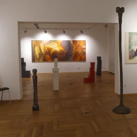 Ein Kunstgalerie mit Holzböden zeigt verschiedene Kunstwerke, darunter abstrakte Gemälde, Skulpturen und einen modernen Stuhl. Beleuchtungskörper hängen von der Decke ab.