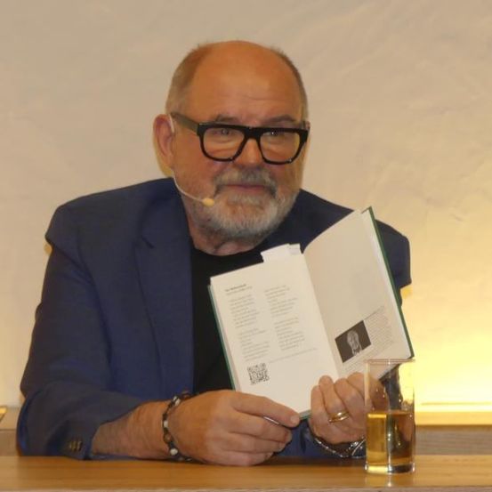 Ein älterer Mann mit Bart und Brille liest ein Buch, während er an einem Tisch mit einem Glas Getränk und einem Mikrofon sitzt.