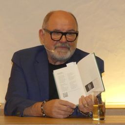 Ein älterer Mann mit Bart und Brille liest ein Buch, während er an einem Tisch mit einem Glas Getränk und einem Mikrofon sitzt.