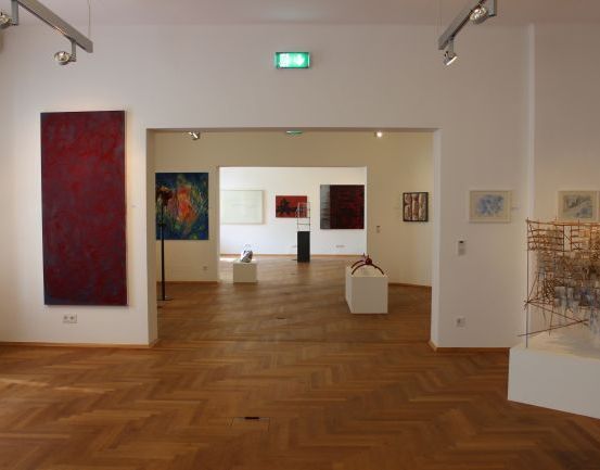 Eine Kunstgalerie mit weißen Wänden und Holzböden. Es gibt Gemälde an den Wänden und Skulpturen auf Podest.