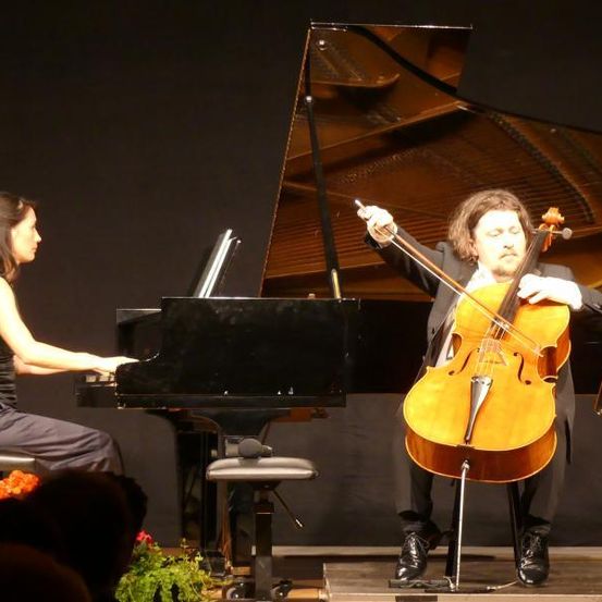 Eine Frau spielt Klavier, während ein Mann Cello auf der Bühne vor einem Publikum spielt.