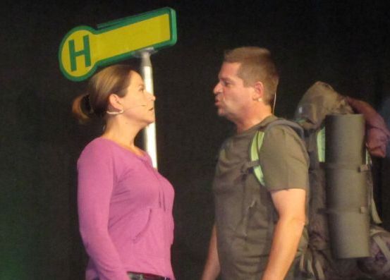 Ein Mann und eine Frau stehen einander in einem schwach beleuchteten Raum gegenüber. Die Frau trägt ein rosa Shirt und ein Mikrofon. Der Mann hat einen Rucksack und sieht ernst aus.