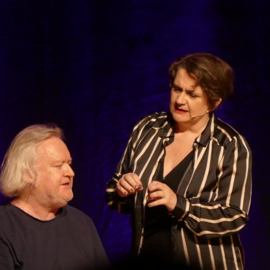 Ein Mann und eine Frau stehen auf einer Bühne unter blauem Licht. Der Mann hat weiße Haare und trägt ein schwarzes T-Shirt. Die Frau trägt ein gestreiftes Oberteil und ein Mikrofon. Sie schaut den Mann an.