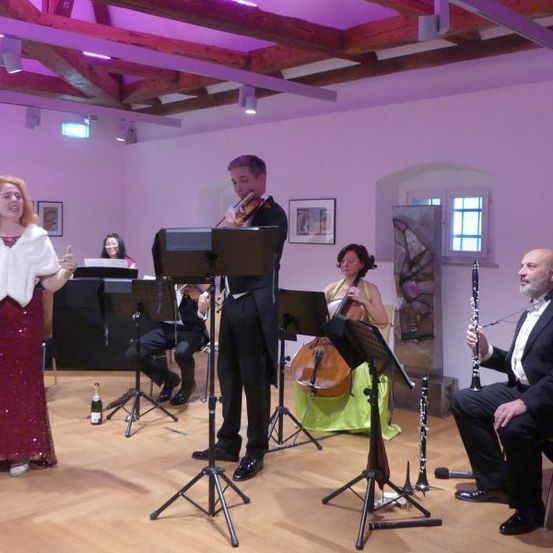 Eine Gruppe von Musikern spielt in einem Raum mit Holzböden. Eine Frau singt, während andere Geige, Cello und Klarinette spielen. Dahinter sitzt eine Pianistin am Klavier. Der Raum hat lila Wände und Holzbalken.