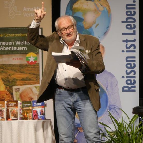 Ein älterer Mann steht auf der Bühne, hält ein Buch in der Hand und liest laut vor einem Hintergrund mit einem Globus und Titeln von Reisebüchern.