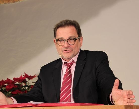 Ein Mann in einem schwarzen Anzug, roter Krawatte und Brille sitzt an einem Rednerpult, gestikuliert mit den Händen und spricht. Dahinter befindet sich eine weiße Wand und eine rote Blumendekoration.