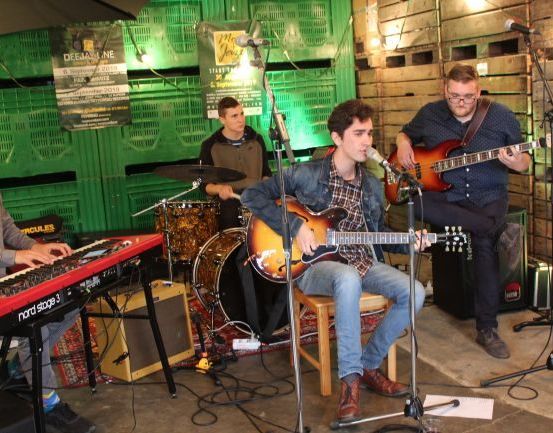 Eine Band aus vier Männern spielt auf der Bühne mit verschiedenen Musikinstrumenten. Ein Mann spielt Klavier, ein anderer Schlagzeug, während zwei weitere Gitarre spielen. Sie stehen vor einem Mikrofon und Lautsprecher.