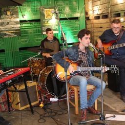 Eine Band aus vier Männern spielt auf der Bühne mit verschiedenen Musikinstrumenten. Ein Mann spielt Klavier, ein anderer Schlagzeug, während zwei weitere Gitarre spielen. Sie stehen vor einem Mikrofon und Lautsprecher.