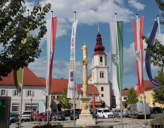 Ein Platz mit Flaggen, einer goldenen Statue, einer Kirche, geparkten Autos und einem Schild vor einem Gebäude.