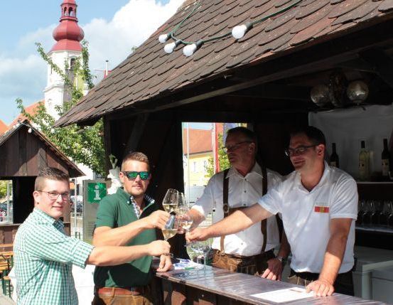 Vier Männer stoßen mit Weingläsern vor einer Bar an. Ein Gebäude mit einer Kirche im Hintergrund.