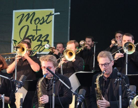 Eine Jazzband tritt auf der Bühne auf, mit Musikern, die Saxophon, Trompete und Posaune spielen, unter einem Schild, das 'Most + Jazz' besagt.