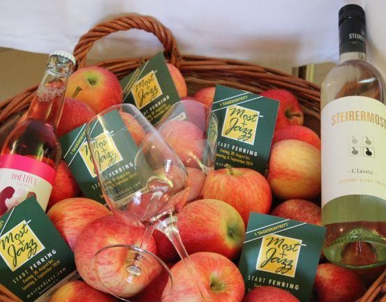 Ein Korb aus Weide gefüllt mit roten Äpfeln, einer Flasche Rotwein und einer Flasche Weißwein. Ein Weinglas steht im Korb, und einige Broschüren sind um die Äpfel herum gelegt.