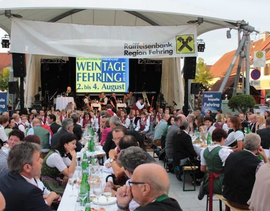 Viele Menschen sitzen an langen Tischen bei einer Veranstaltung im Freien, wahrscheinlich einem Festival. Eine Bühne mit einer Band ist sichtbar, und ein Banner trägt den Text 'Weintage Fehring 2019'.