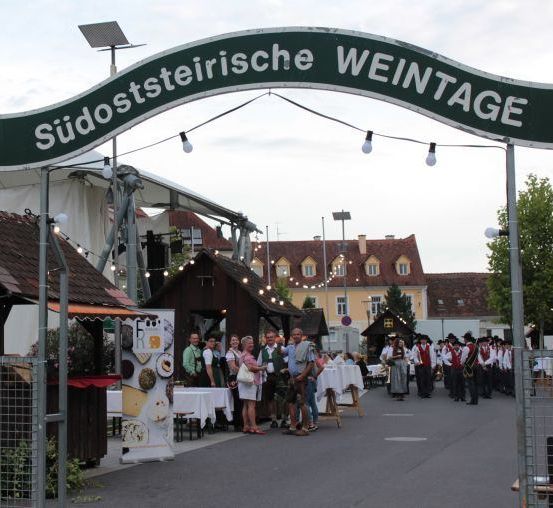 Men und Frauen in traditioneller Kleidung stehen unter einem Bogen mit der Aufschrift 'Sudoststeiriche Weintage', in der Nähe von Essensständen und einer Musikkapelle auf einem Stadtplatz.