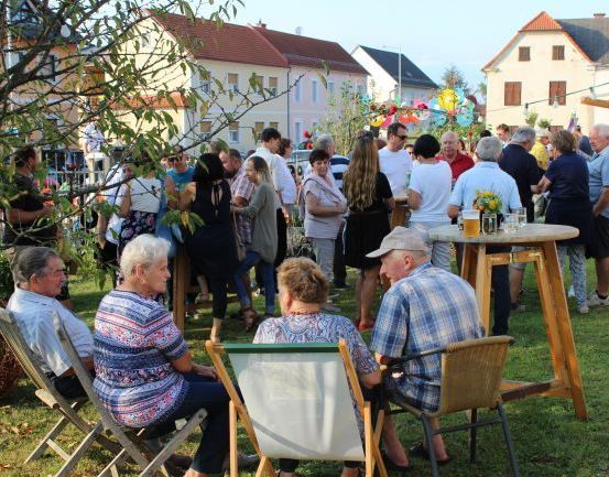 Eine Gruppe älterer Menschen trifft sich in einem Garten, einige sitzen in Stühlen, andere stehen. Ein Tisch hält Getränke, und Häuser sind im Hintergrund zu sehen.
