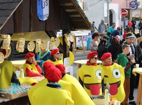 Eine Menschenmenge in Kostümen versammelt sich auf einem Straßenfest. Einige tragen Smiley-Gesichtskostüme, während andere rote Mützen tragen. Tische mit Gläsern und Essen sind aufgestellt.