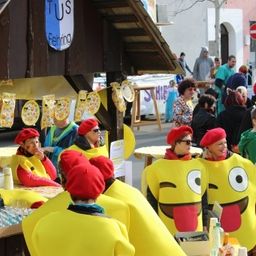 Eine Menschenmenge in Kostümen versammelt sich auf einem Straßenfest. Einige tragen Smiley-Gesichtskostüme, während andere rote Mützen tragen. Tische mit Gläsern und Essen sind aufgestellt.