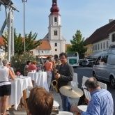 Ein Straßenmusiker spielt Saxophon vor einem Outdoor-Restaurant mit Tischen und Stühlen, während eine Menge zuschaut. Im Hintergrund steht eine Kirche mit Turm.