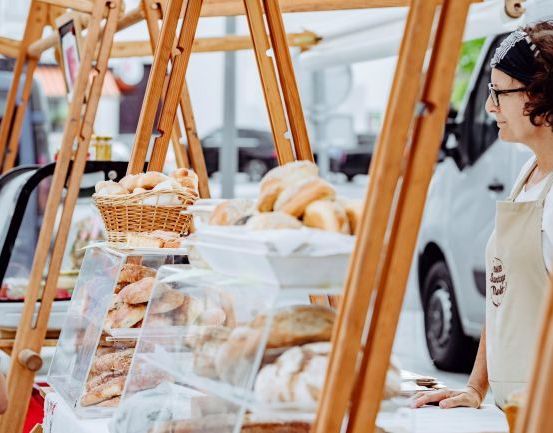 Eine Frau in einer Schürze steht hinter einer Ausstellung von Backwaren auf einem Outdoor-Markt, mit einem Korb voller Brot im Hintergrund und einem weißen Van, der in der Nähe geparkt ist.
