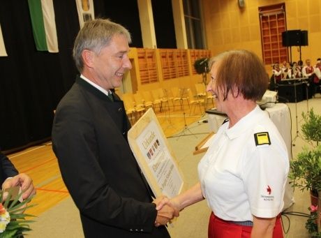 Ein Mann und eine Frau, formal gekleidet, schütteln sich die Hände. Der Mann hält eine Urkunde, und die Frau lächelt. Hinter ihnen sitzt eine Gruppe von Menschen.