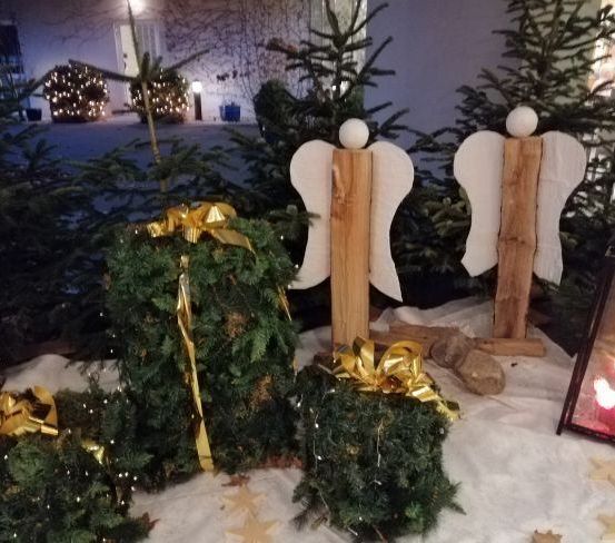 Dekorierte Weihnachtsbäume und zwei hölzerne Engelfiguren mit weißen Flügeln stehen auf einem weißen Tuch. Ein Laterne und gelbe Sterne sind auf dem Boden.
