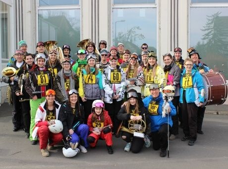 Eine Gruppe von Menschen in Winterkleidung, Helmen und Nummernschildern posiert für ein Foto vor einem Gebäude. Sie halten Musikinstrumente.