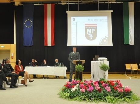 Ein Mann in Uniform hält eine Rede an einem Podium mit einer Projektionsleinwand, auf der 'MMP' steht. Viele Menschen sitzen an einem Tisch, und Flaggen hängen an der Wand.