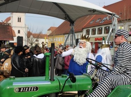 Eine Gruppe von Menschen in Kostümen versammelt sich um einen grünen Traktor mit einem großen Baldachin. Eine Person, die als König verkleidet ist, sitzt im Traktor, während eine andere in gestreifter Kleidung zuschaut. Dahinter ist ein Gebäude mit einem roten Dach und mehreren Fenstern zu sehen.