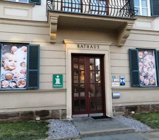 Der Eingang eines Gebäudes mit der Aufschrift Rathaus, mit einer braunen Tür und Fenstern, die mit Babybildern geschmückt sind.