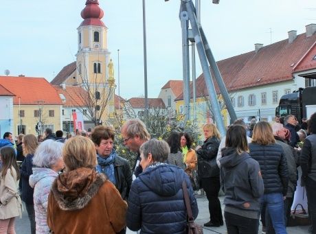 Eine Gruppe von Menschen versammelt sich auf einem Stadtplatz, mit einem Kirchturm im Hintergrund. Einige tragen Wintermäntel und Schals.