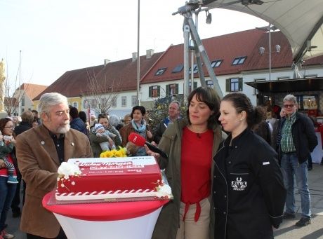Ein Mann hält einen Kuchen, während zwei Frauen zuhören, eine mit einem Mikrofon, vor einer Menge. Dahinter steht ein Gebäude mit einem roten Dach.