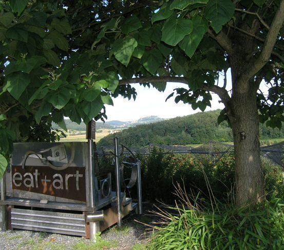 Eine Außenstruktur mit 'eat+art' darauf steht unter einem großen Baum. Der Hintergrund zeigt Hügel, Bäume und Pflanzen, mit einem Blick auf eine entfernte Bergkette.