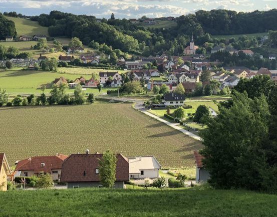 Luftaufnahme eines ländlichen Dorfes mit Häusern, einer Kirche und umliegenden Feldern und Bäumen.