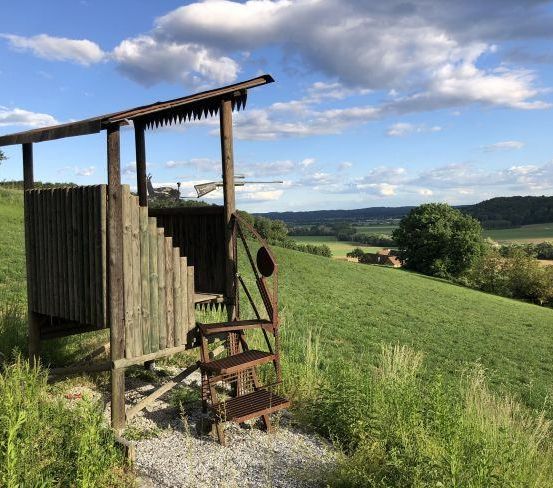 Ein alter Holzstand auf einem grasbewachsenen Hügel mit einem Gewehr und einer Leiter. Bäume und eine entfernte Aussicht auf ein Tal.
