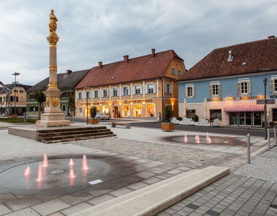 Ein Stadtplatz mit einem Brunnen, einer Statue und Gebäuden mit Geschäften und Cafés.