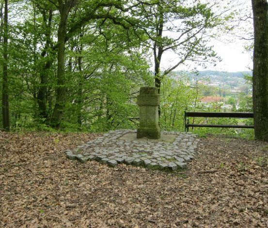 Ein Steinmonument steht in der Mitte eines kreisförmigen Steinpfades, umgeben von Bäumen und Laub. Rechts ist eine Bank zu sehen.