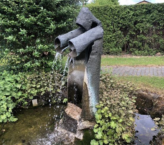 Ein Betonbrunnen mit Wasser, das aus Rohren fließt, steht in einem Garten, umgeben von grünen Pflanzen und Bäumen. Das Wasser spritzt in einen Teich.