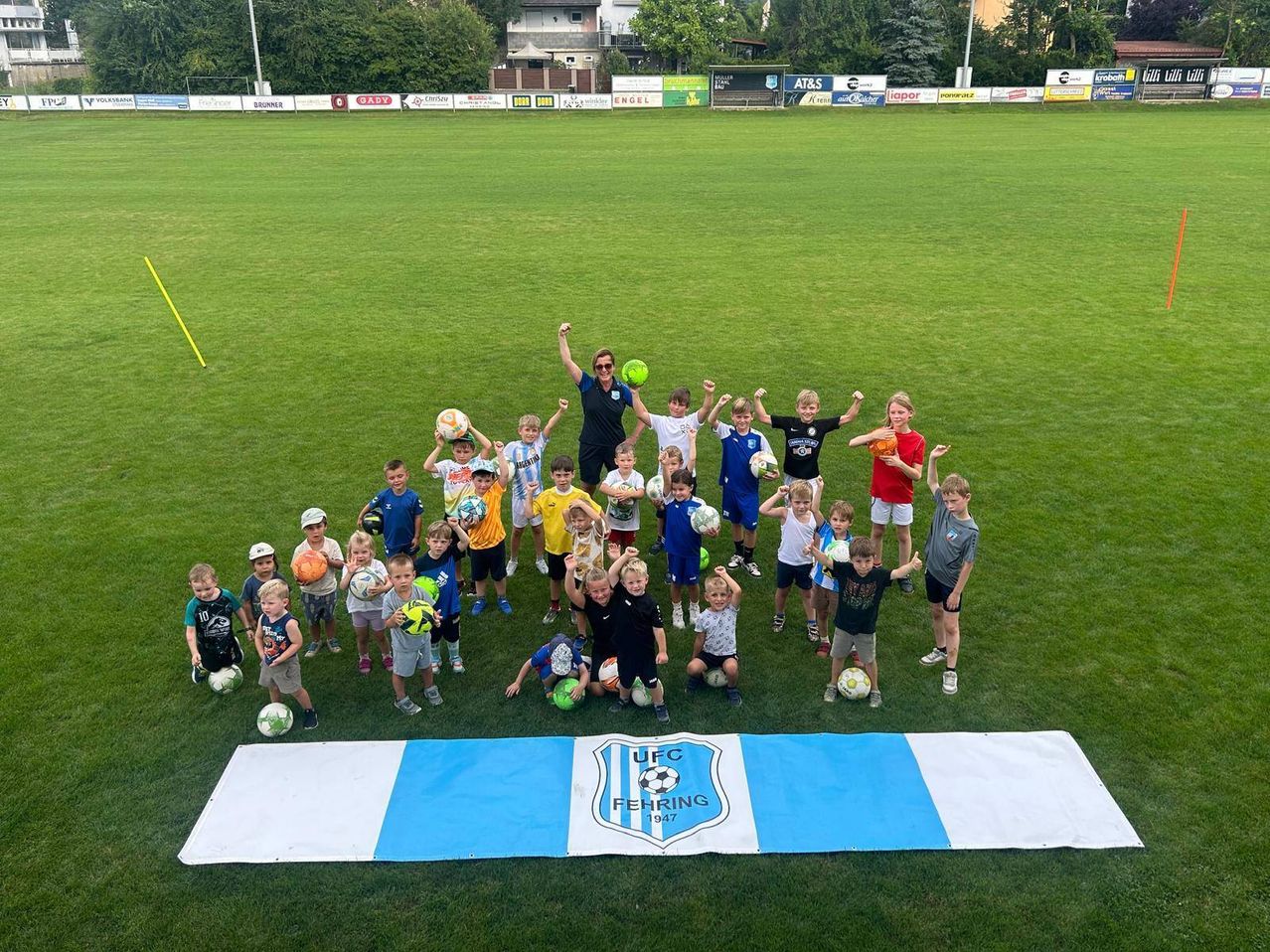 Eine Gruppe von Kindern und Erwachsenen auf einem Fußballfeld mit blauen und weißen Bannern im Vordergrund. Sie halten Fußballbälle und feiern.