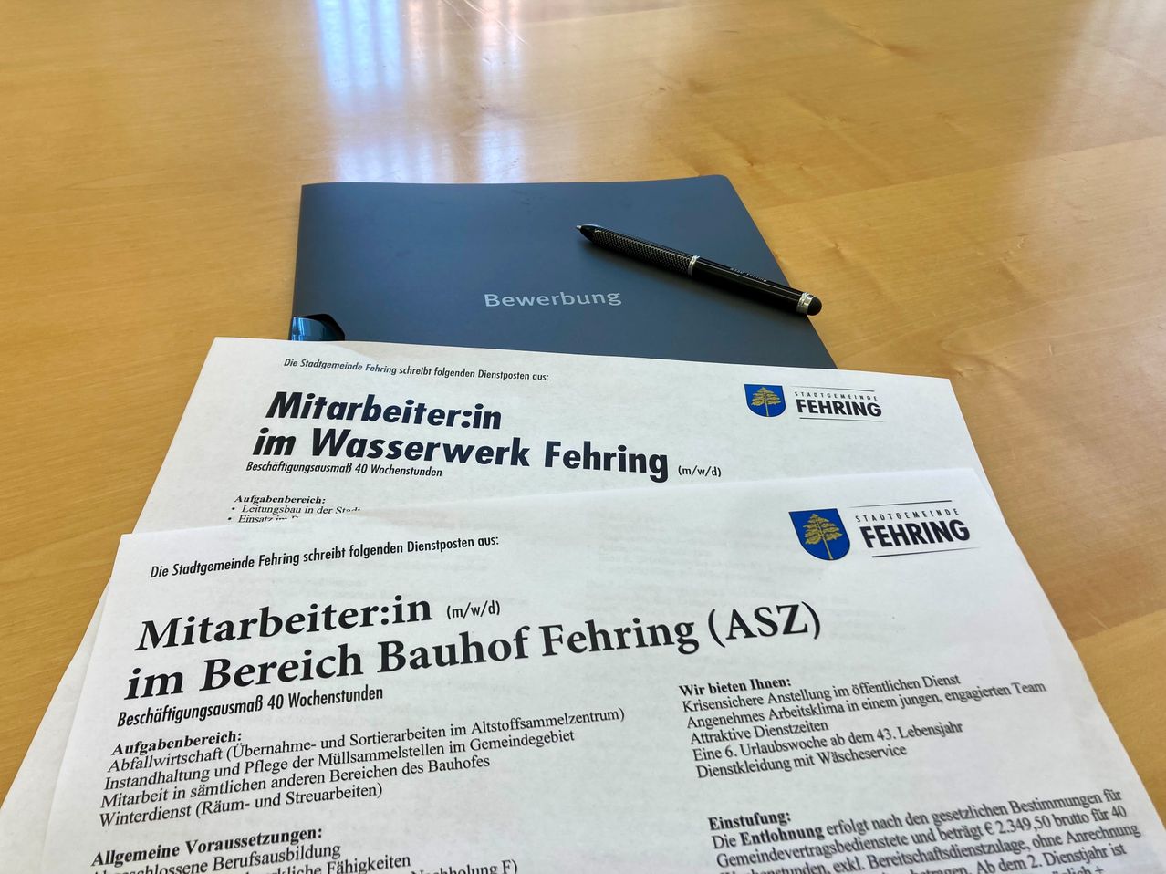 Ein Bewerbungsschreiben der Gemeinde Fehring liegt auf einem Holztisch mit einer blauen Mappe, die Bewerbung beschriftet ist. Die Bewerbung umfasst eine Stellenbeschreibung für eine Teilzeitstelle in den Wasserwerken von Fehring. Ein Stift ruht auf der Mappe.