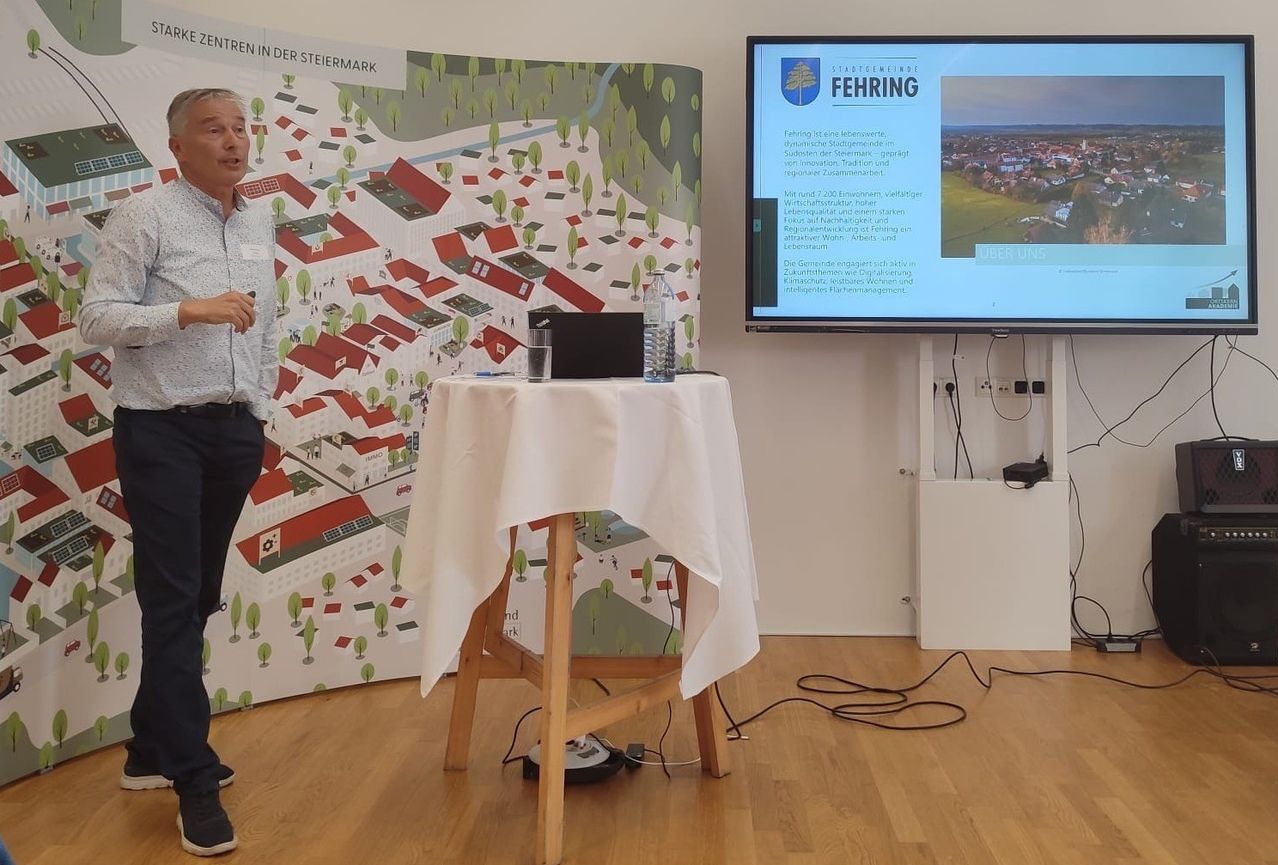 Ein Mann steht vor einer Karte und hält eine Präsentation. Auf einem Tisch befinden sich ein Laptop, Wasserflaschen und ein Becher. Ein Monitor zeigt eine Präsentation mit Text an. Der Boden ist aus Holz und die Wände haben eine Wandmalerei.