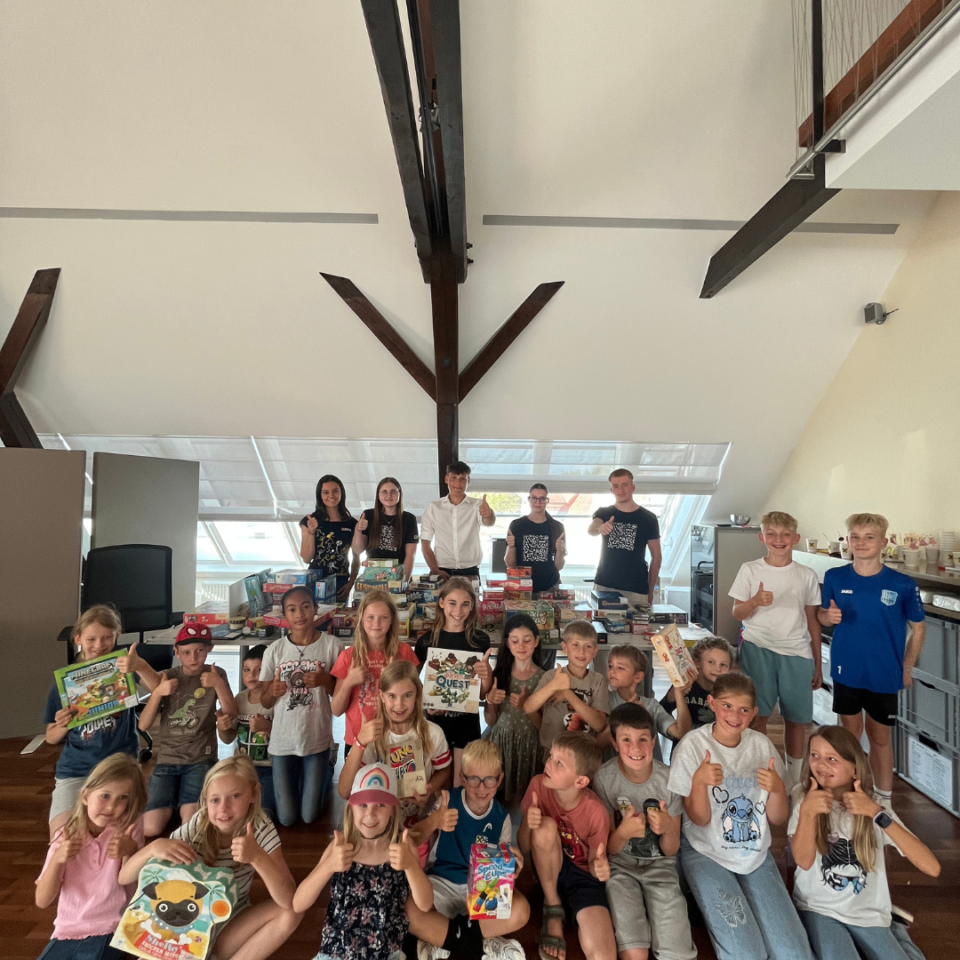Eine Gruppe von Kindern und Erwachsenen versammelt sich in einem Raum, lächelt und hält Geschenke, mit einem Schreibtisch und Lagerboxen im Hintergrund.