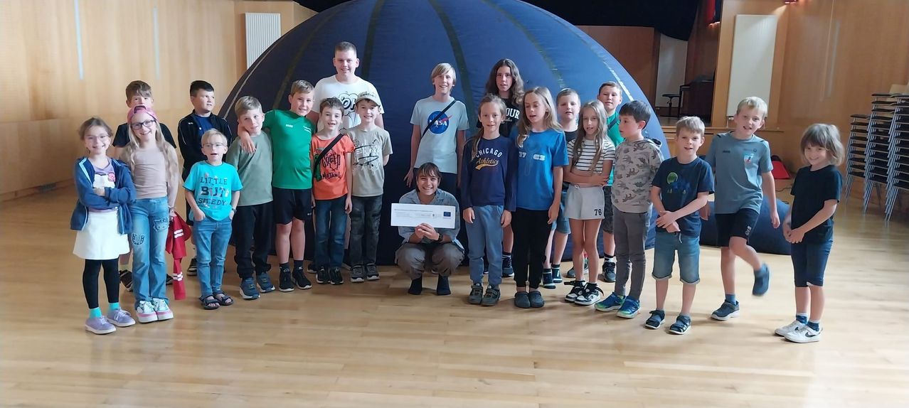 Eine Gruppe von Kindern steht vor einem blauen aufblasbaren Planetarium und hält ein Zertifikat. Sie alle lächeln für ein Foto.