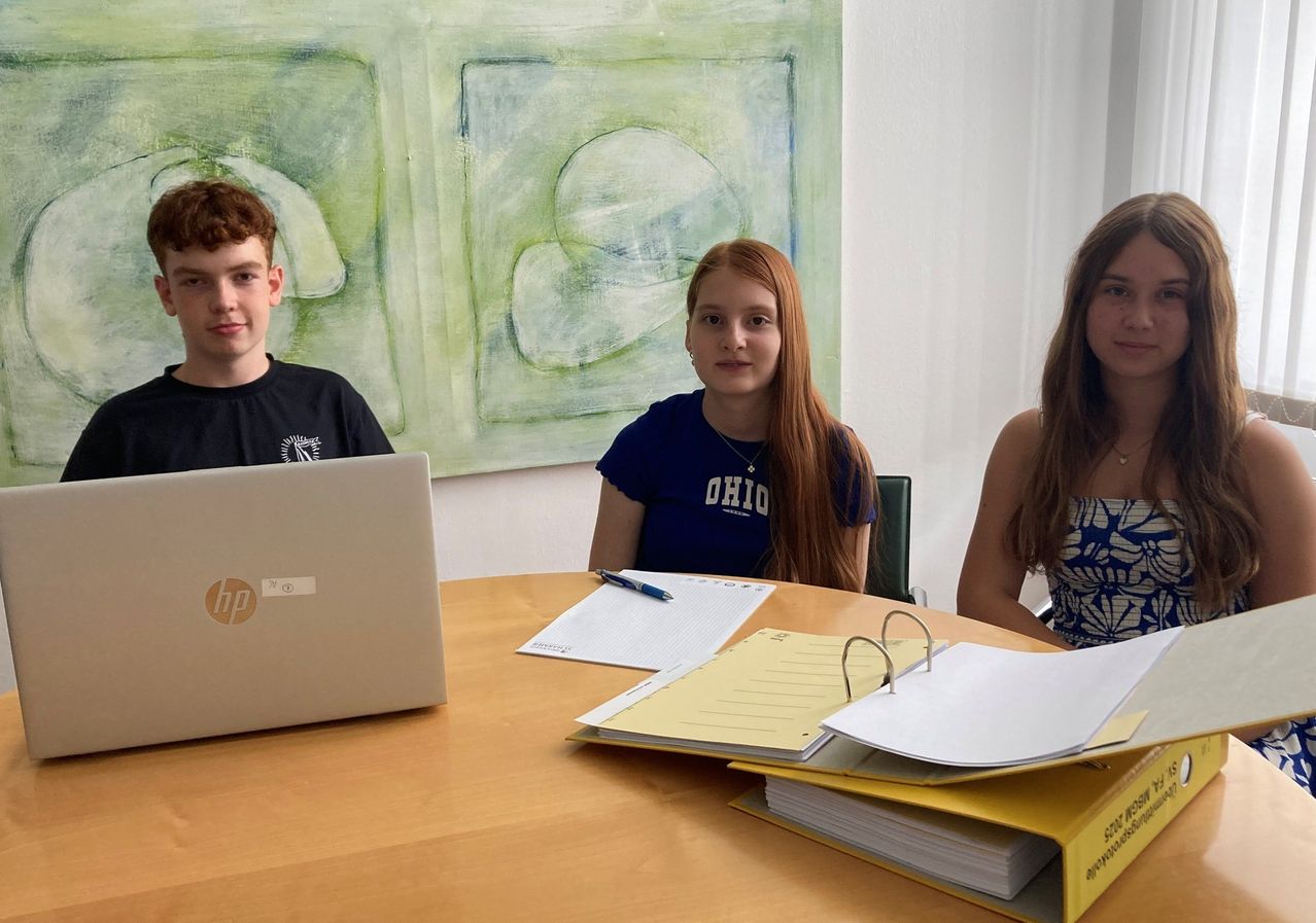 Drei junge Personen sitzen um einen Tisch herum und schauen in die Kamera. Ein junger Mann ist links, eine junge Frau in einem blauen T-Shirt in der Mitte und eine weitere junge Frau rechts. Ein Laptop und Papiere sind auf dem Tisch.