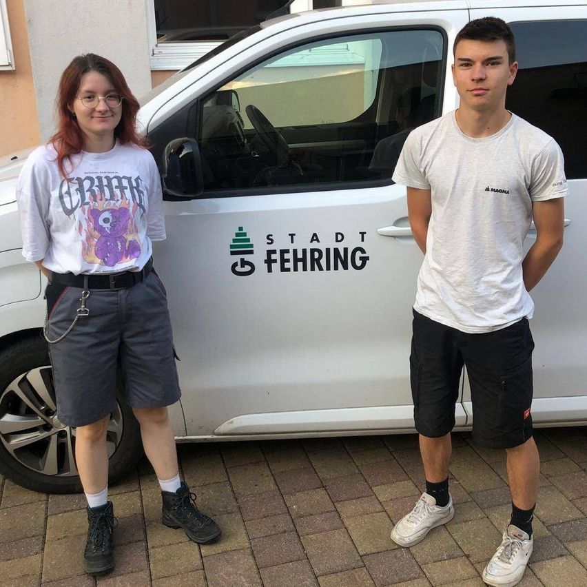 Zwei Personen stehen vor einem weißen Van mit der Aufschrift Stadt Fehing an der Seite. Die Frau trägt eine Brille und ein weißes T-Shirt mit einem grafischen Design, während der Mann ein graues T-Shirt trägt. Sie lächeln beide und scheinen für ein Foto zu posieren.