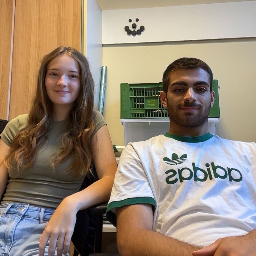 Eine junge Frau und ein Mann sitzen zusammen in einem Büro und lächeln für ein Foto. Die Frau trägt ein grünes T-Shirt und Jeansshorts, während der Mann ein weißes Adidas-Shirt trägt. Hinter ihnen steht ein Holzschrank und eine grüne Aufbewahrungsbox.