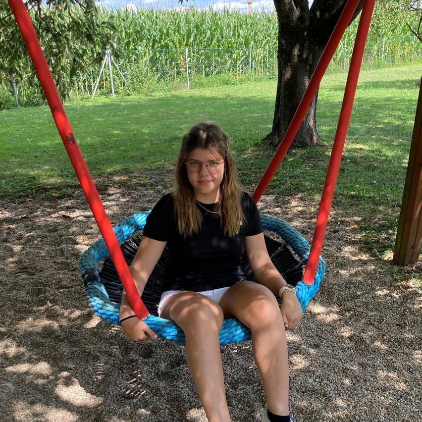 Eine Frau in einem schwarzen T-Shirt und weißen Shorts sitzt auf einem blauen Schaukel mit roten Griffen in einem Außenbereich mit einem Baum und einem Maisfeld im Hintergrund.