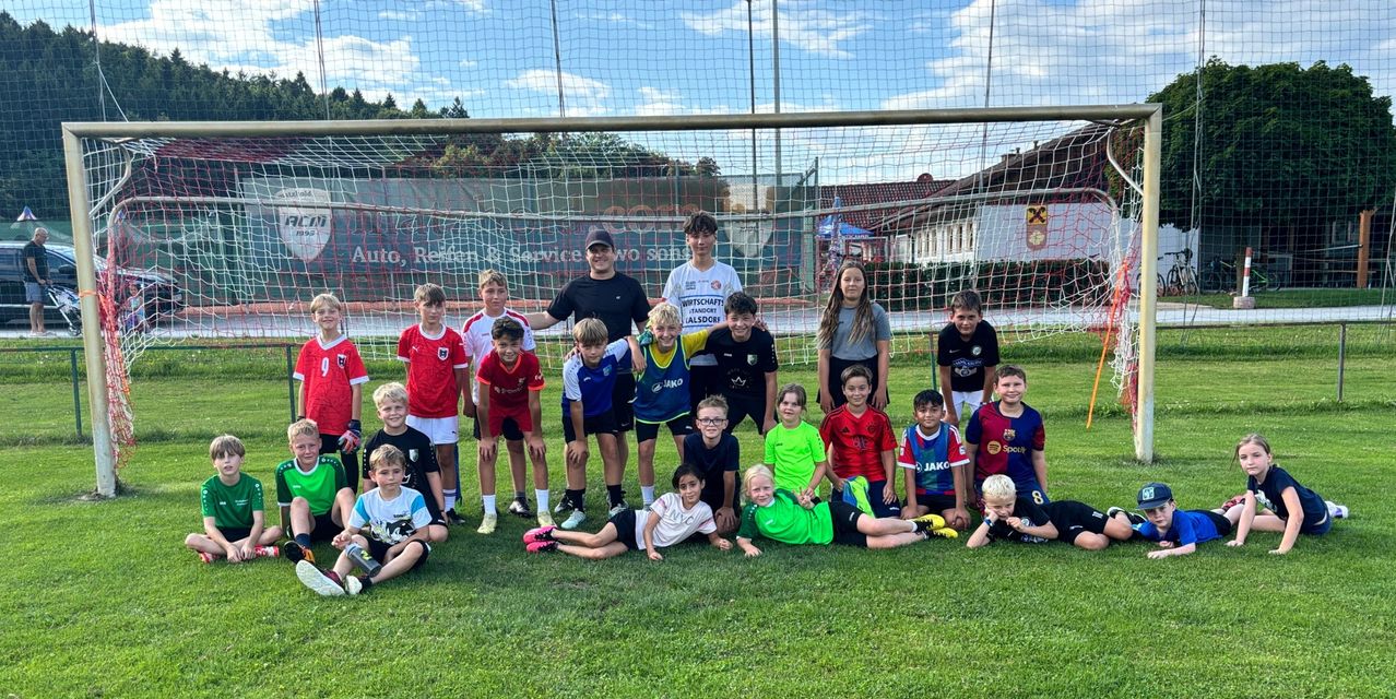 Eine Jugendfußballmannschaft, bestehend aus Kindern und Erwachsenen, posiert für ein Gruppenfoto vor einem Fußballtor. Sie tragen verschiedenfarbige Trikots, und einige halten Fußballbälle.