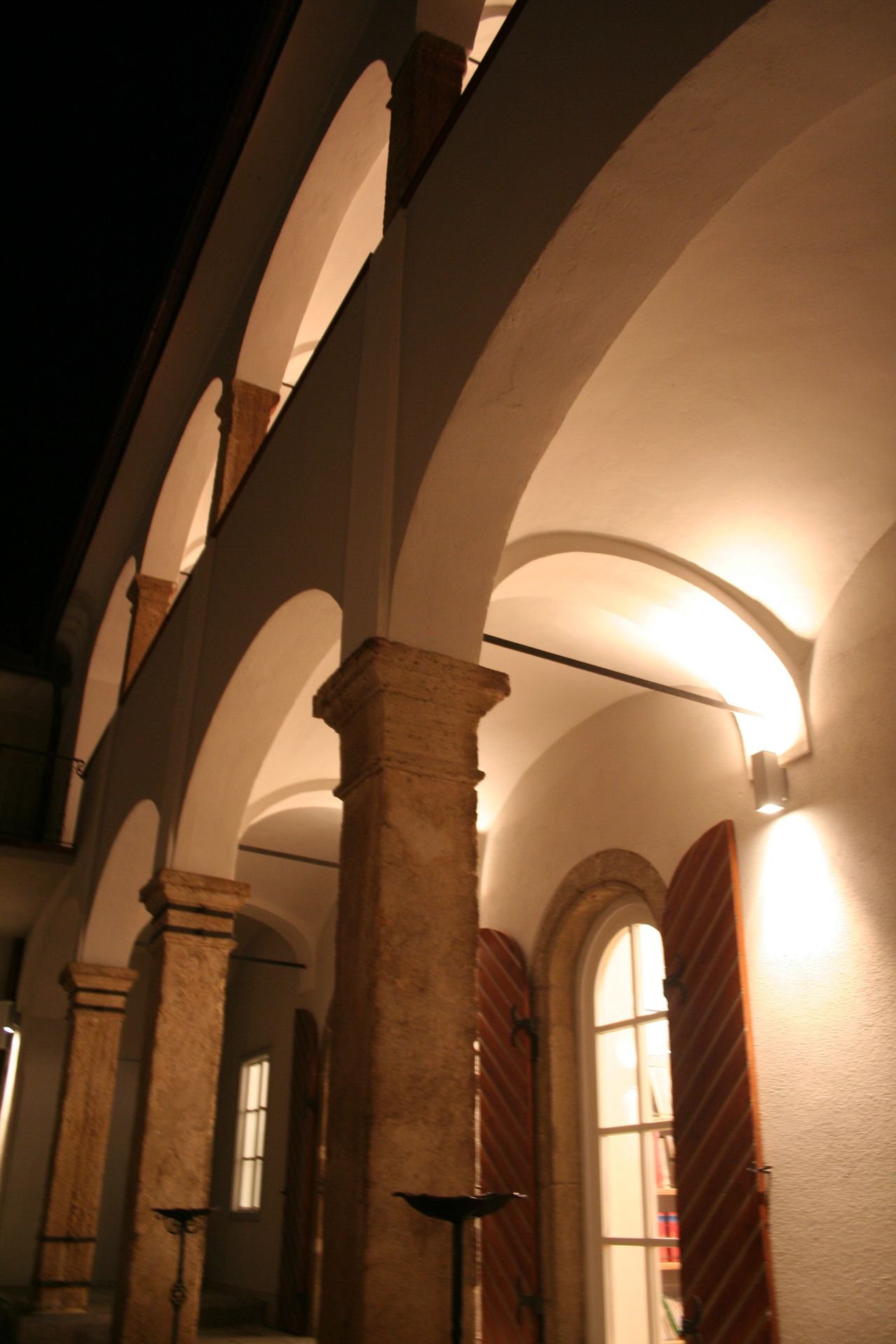 Eine Innenansicht eines Gebäudes mit gewölbten Decken, Steinsäulen und geschlossenen Holzläden, bei Nacht von warmen Lichtern beleuchtet.