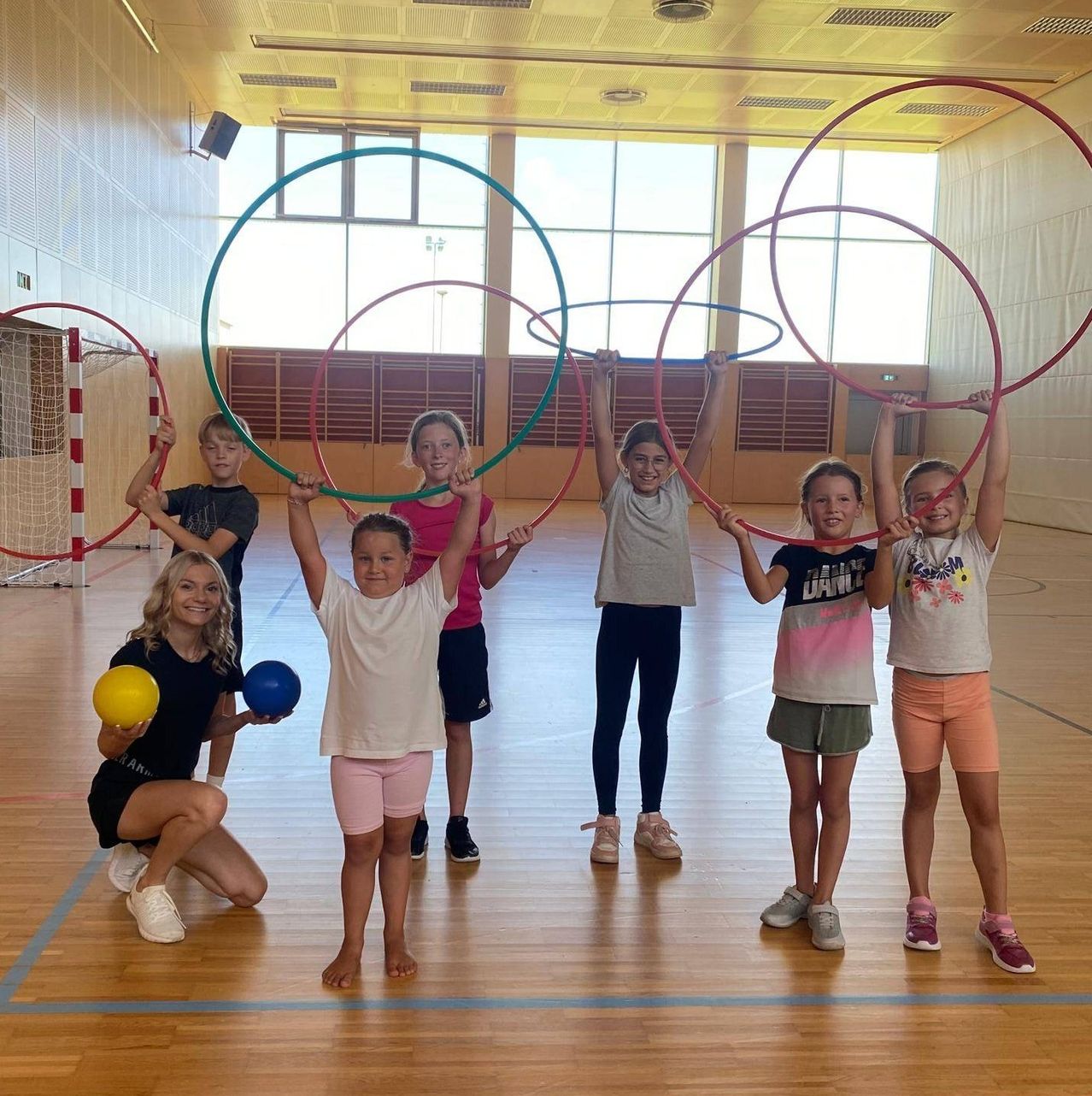 Mehrere Kinder und eine erwachsene Frau sind in einer Turnhalle und halten Hula-Hoops. Sie lächeln und posieren für ein Foto.