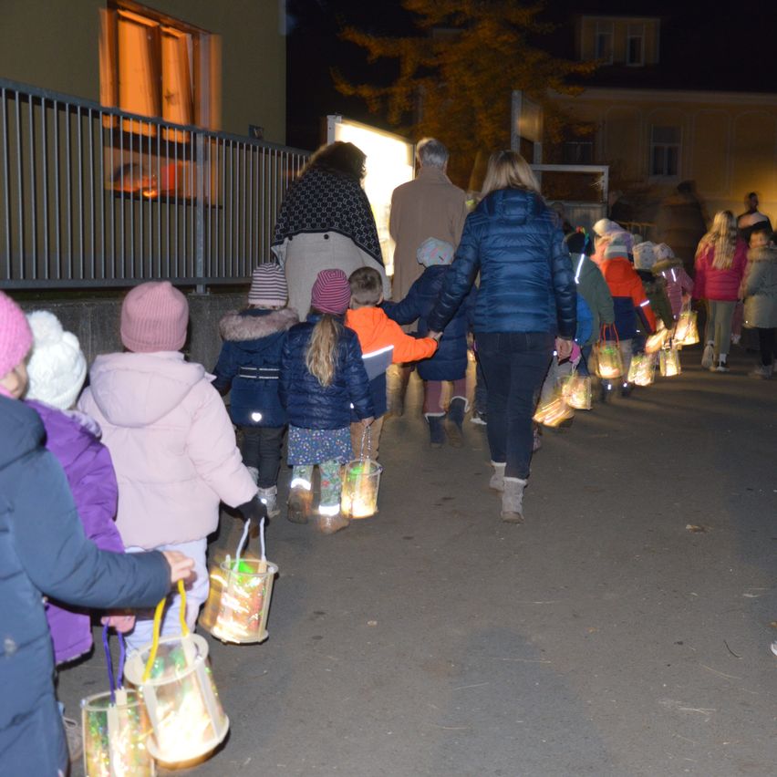 Eine Gruppe von Kindern mit Laternen geht mit einer Frau und einem Mann an der Spitze einen Bürgersteig entlang. Ein Zaun und ein Gebäude sind im Hintergrund.